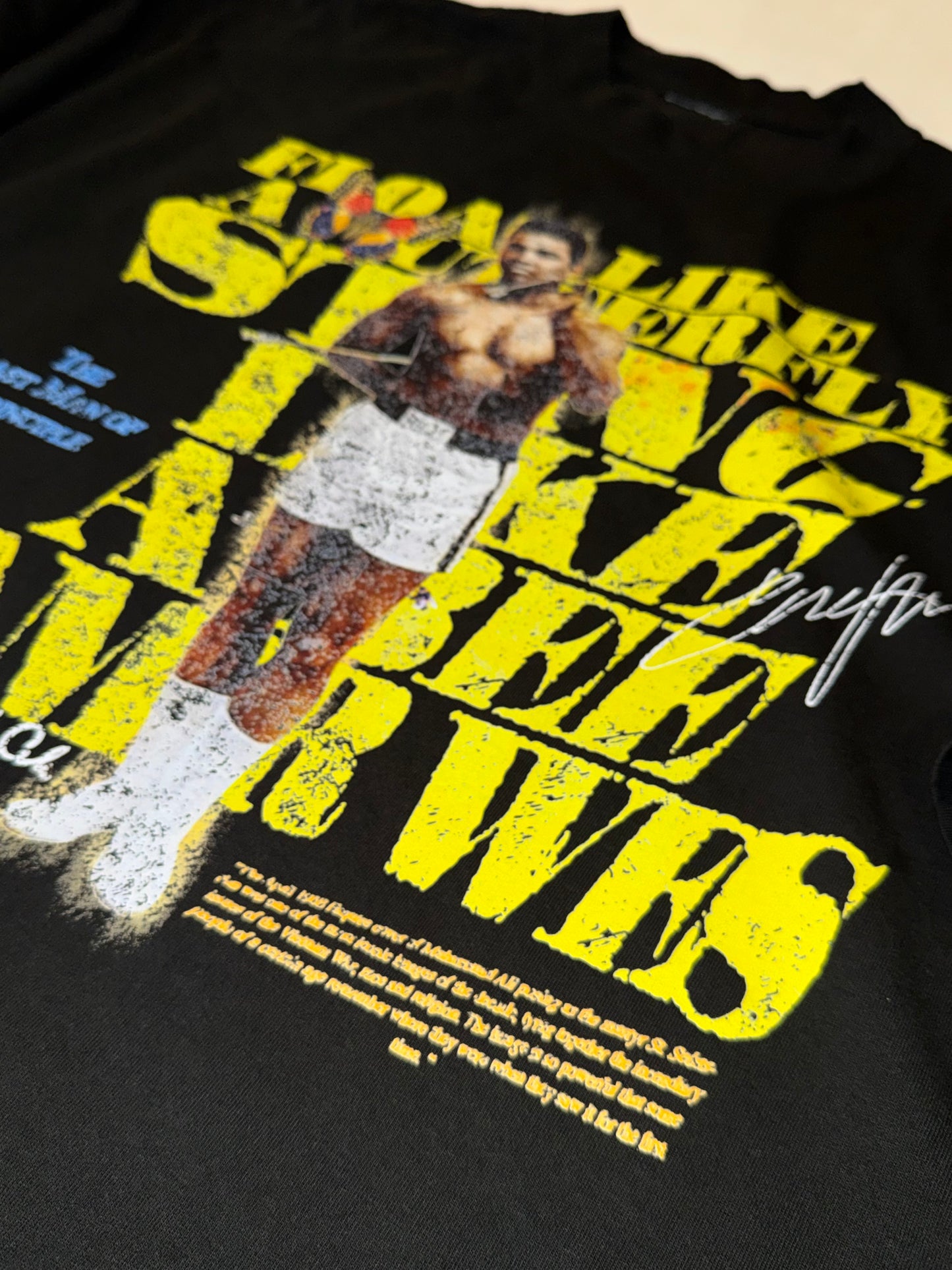 A black short-sleeve graphic t-shirt laid flat on a white background. The shirt features a large front print with yellow text reading 'FLOAT LIKE A BUTTERFLY STING LIKE A BEE LIKE A BEE' and a central image of Amir Wes.