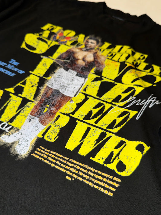 A black short-sleeve graphic t-shirt laid flat on a white background. The shirt features a large front print with yellow text reading 'FLOAT LIKE A BUTTERFLY STING LIKE A BEE LIKE A BEE' and a central image of Amir Wes.
